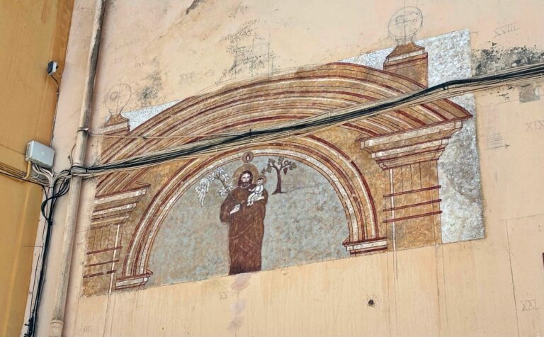 Faded wall mural showing a saint beneath a decorative arched arch on a yellow wall, with tangled wires and an electrical box on the left.