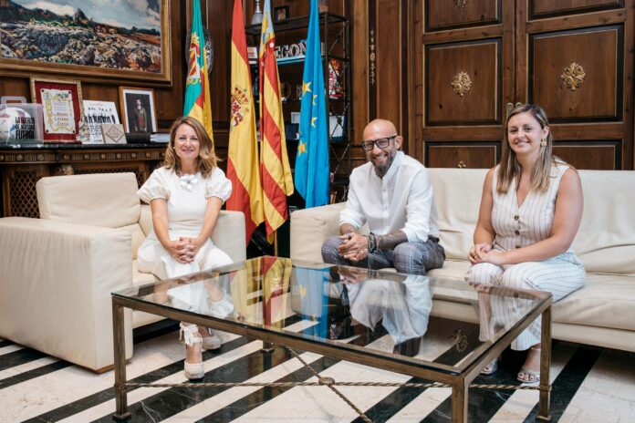 Three adults seated on cream sofas in a wood-paneled room with flags behind them and a glass coffee table in the foreground.