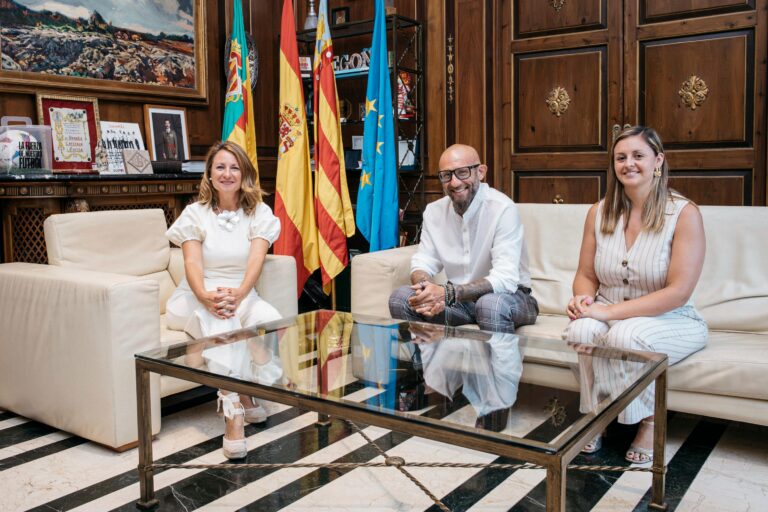 Three adults seated on cream sofas in a wood-paneled room with flags behind them and a glass coffee table in the foreground.