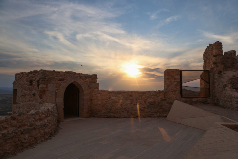 Ancient stone ruins at sunset with an arched doorway and rocky walls, a warm sun low on the horizon.