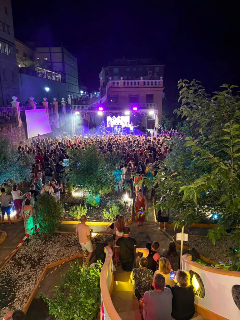 Nighttime outdoor concert with a large crowd facing a brightly lit stage, surrounded by buildings and greenery.