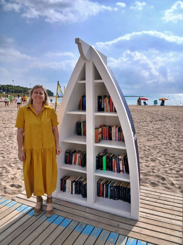 Woman in a yellow dress stands beside a large white boat-shaped bookshelf on a sandy beach with the ocean in the background.