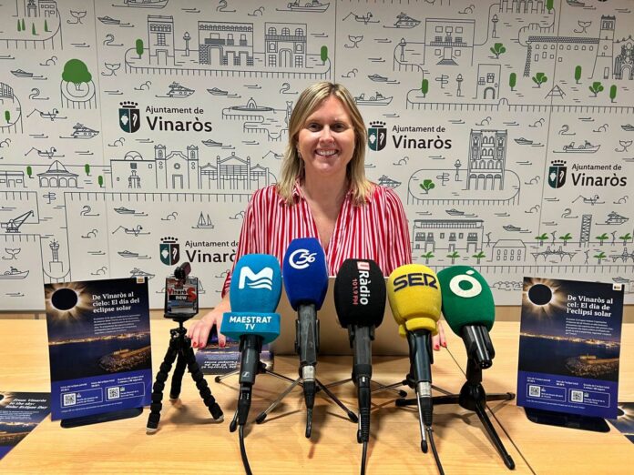 Smiling woman in a red-striped blouse stands behind multiple microphones at a press conference, with a city backdrop reading 'Ajuntament de Vinaròs'.