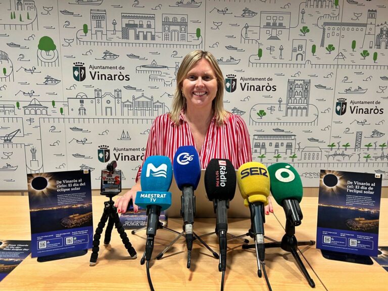 Smiling woman in a red-striped blouse stands behind multiple microphones at a press conference, with a city backdrop reading 'Ajuntament de Vinaròs'.