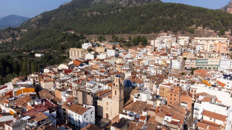 Aerial view of a dense town at the base of forested hills with orange-tiled rooftops and pastel buildings.