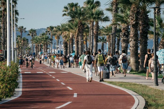 Crowded seaside promenade with pedestrians and cyclists along a palm-tree lined bike path as the sun shines.