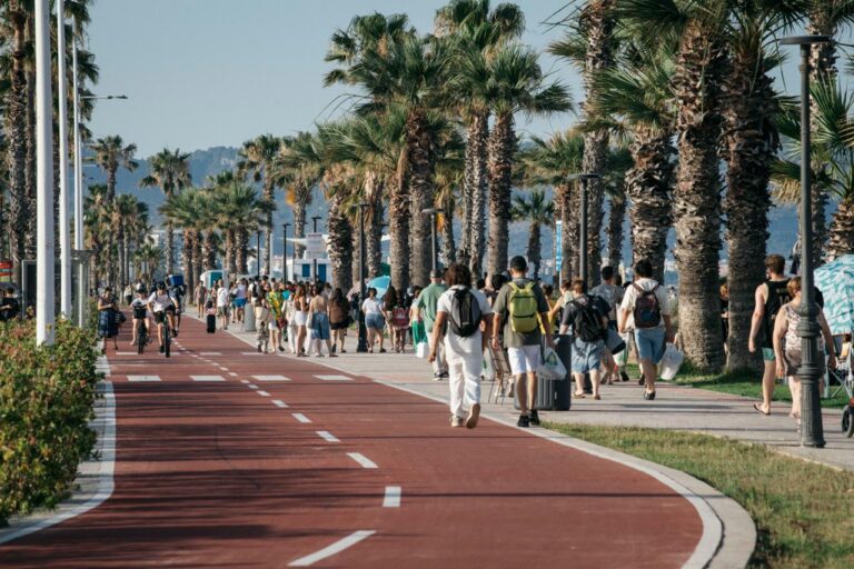 Crowded seaside promenade with pedestrians and cyclists along a palm-tree lined bike path as the sun shines.