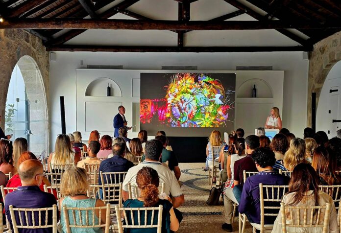Audience watches a presentation in a hall with a large colorful abstract artwork on the screen at the front.