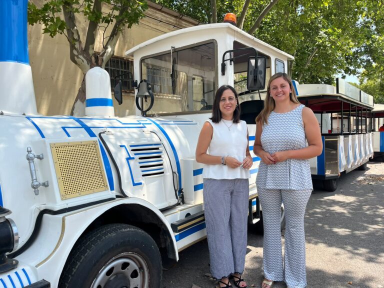 Two women pose beside a white and blue locomotive with open passenger cars outdoors, smiling on a sunny day.