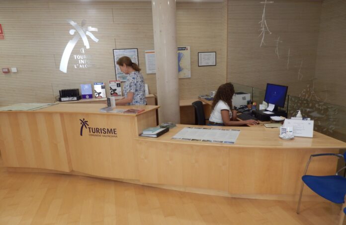 Tourist information desk with two staff members assisting visitors at a wooden counter, brochures and maps on display, branding in Valencia tourism.