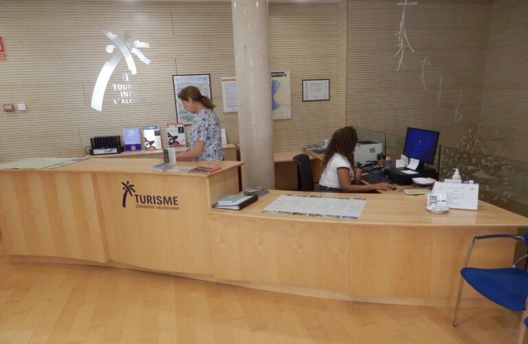 Tourist information desk with two staff members assisting visitors at a wooden counter, brochures and maps on display, branding in Valencia tourism.