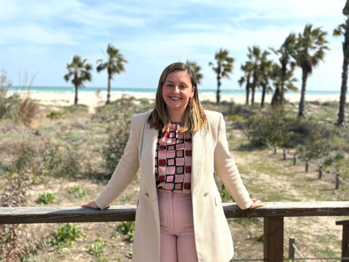 Smiling woman in a beige coat standing at a wooden railing with palm trees and a beach in the background