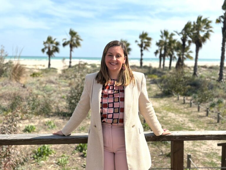 Smiling woman in a beige coat standing at a wooden railing with palm trees and a beach in the background