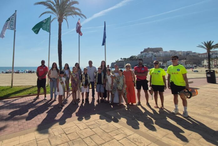 Group of people posing on a sunny beachside promenade with flags, palm trees, and a coastal town in the background.