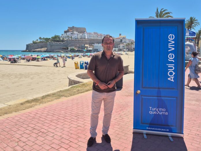 Man in brown polo shirt and light pants stands on a red brick promenade by a blue promotional kiosk on a sunny beach, with umbrellas and a historic fortress behind him.