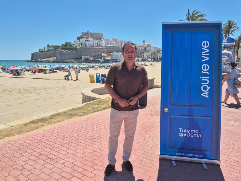 Man in brown polo shirt and light pants stands on a red brick promenade by a blue promotional kiosk on a sunny beach, with umbrellas and a historic fortress behind him.