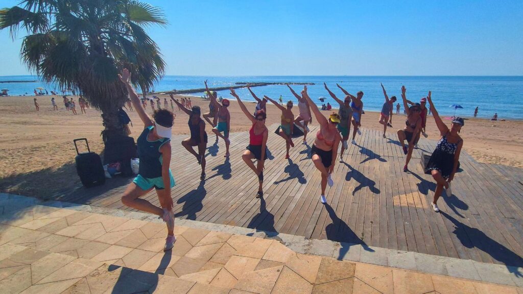 Group of people doing synchronized balance pose on a wooden boardwalk by the beach under a sunny sky, palm tree nearby.