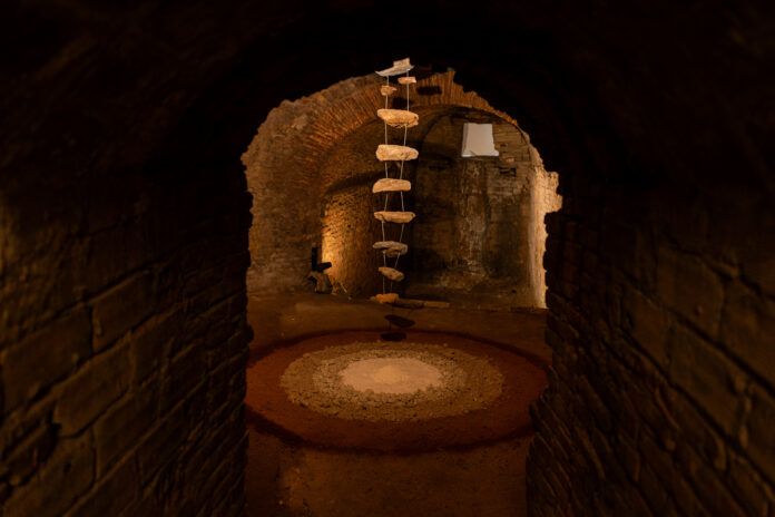 Dark underground brick tunnel with a hanging rope ladder made of stone steps and a circular rubble-filled pit on the floor in the foreground.