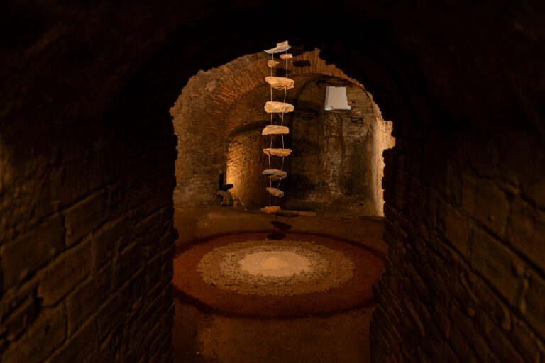 Dark underground brick tunnel with a hanging rope ladder made of stone steps and a circular rubble-filled pit on the floor in the foreground.