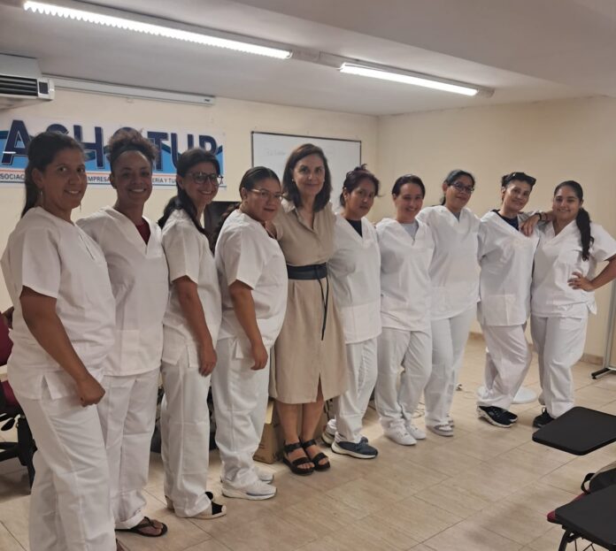 Line of healthcare workers in white scrubs posing for a group photo in a training room; a woman in a beige dress stands center among them.