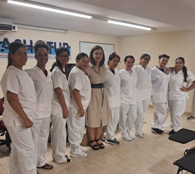 Line of healthcare workers in white scrubs posing for a group photo in a training room; a woman in a beige dress stands center among them.