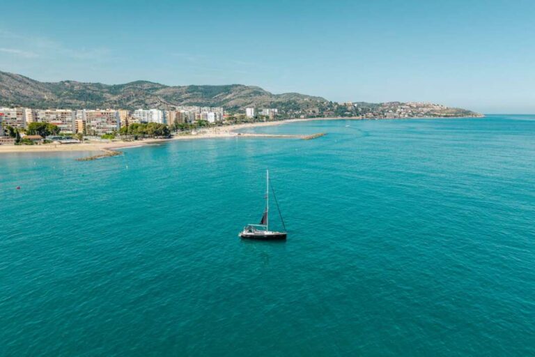 A calm turquoise sea with a single sailboat centered, and a coastal town with buildings and hills in the background under a clear blue sky