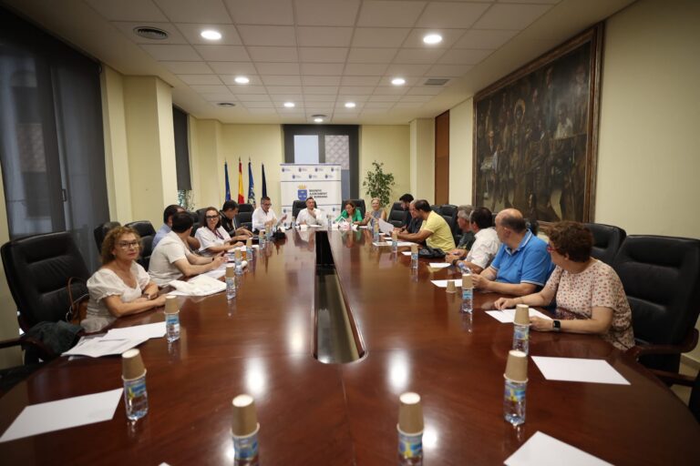 Long conference table with many people in a meeting room, water bottles and papers, banner and flags in the background.