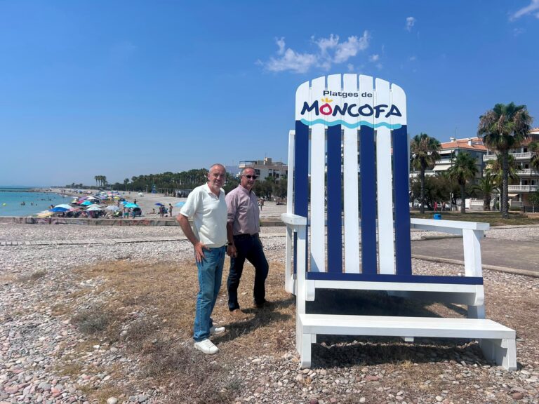 Two men stand beside a tall white-and-blue striped beach sign on a pebbled shore, with umbrellas and the sea in the background.
