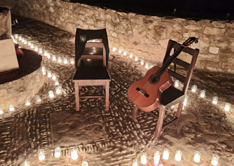Two wooden chairs sit in a candlelit stone courtyard at night, with a classical guitar resting on one chair.