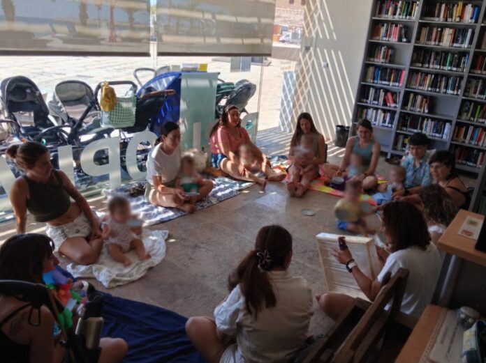 Parents and caregivers sit in a circle on blankets with babies in a library reading area, with bookshelves along one wall and strollers outside the window.