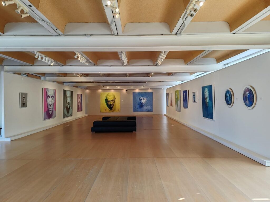 Wide view of a contemporary art gallery with colorful portraits on white walls and black benches in the center.