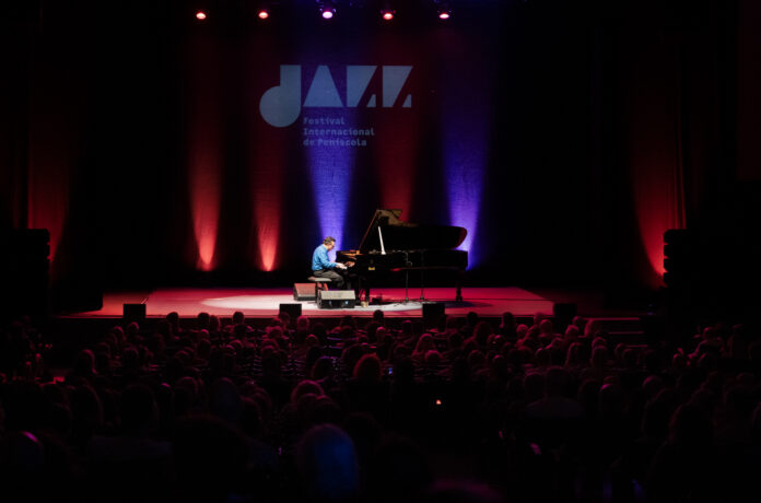Pianist in a blue shirt performing on a grand piano under colorful red and blue stage lights with an audience in silhouette.