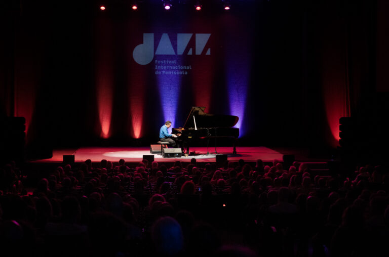Pianist in a blue shirt performing on a grand piano under colorful red and blue stage lights with an audience in silhouette.