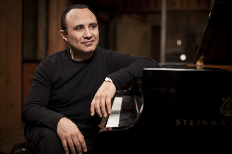 Man in a black sweater seated at a grand piano, looking to the left in a warmly lit room.