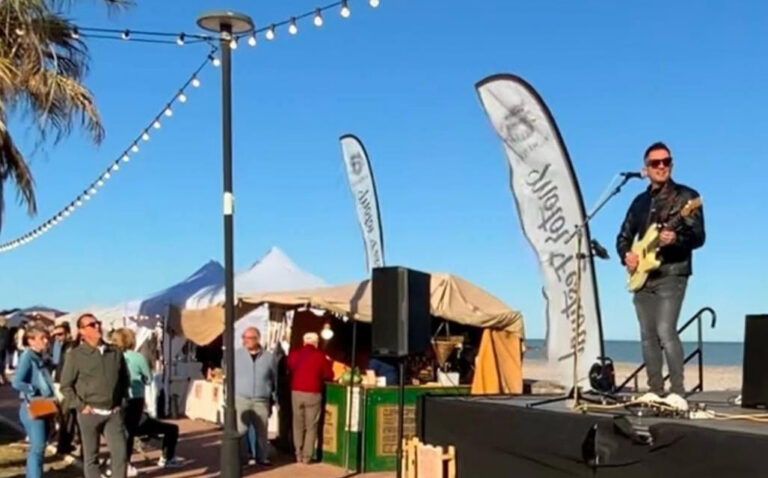 A guitarist wearing sunglasses performs on a small stage at a sunny beach market, with string lights overhead, white tents and market stalls along the sand, and people browsing in the background with the ocean visible.