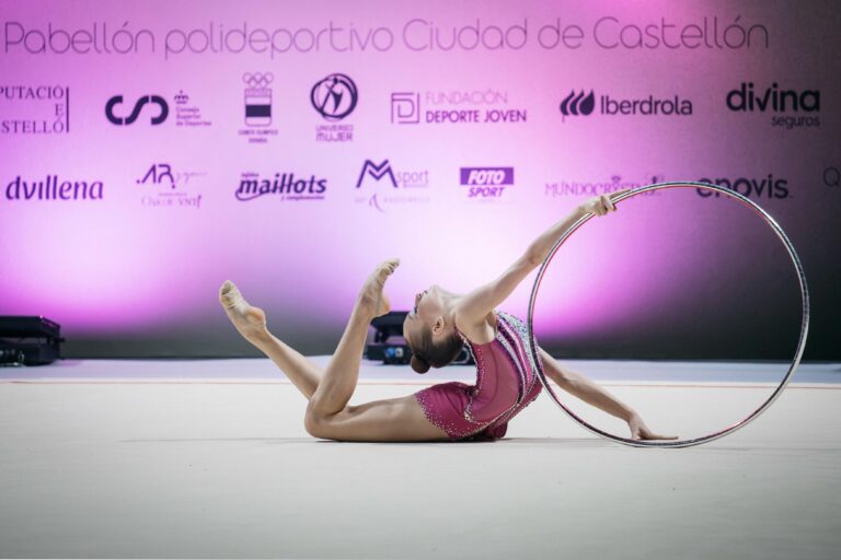 Two rhythmic gymnasts perform with a hoop on a pink-lit competition stage with sponsor logos in the background on the wall behind them.