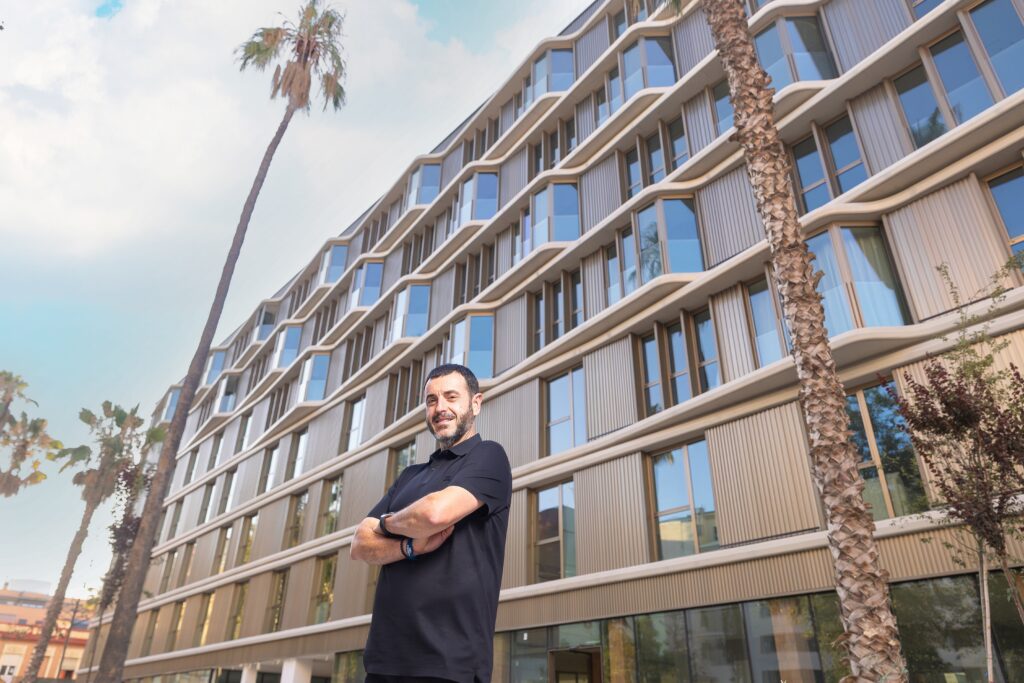 Man in a black polo with arms crossed stands outside a modern glass-and-tiber facade building with palm trees nearby.