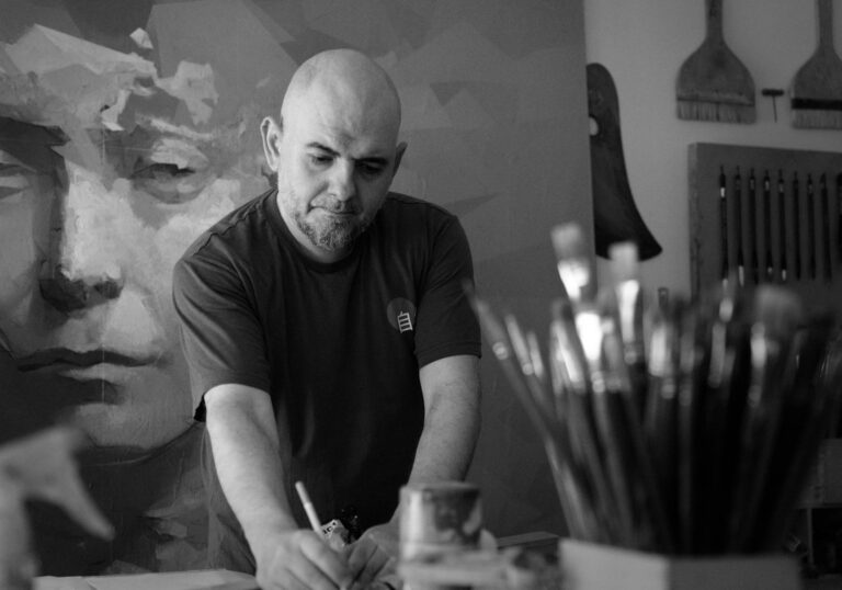 Bald man wearing a dark shirt focused on painting at a cluttered art studio with brushes in the foreground.