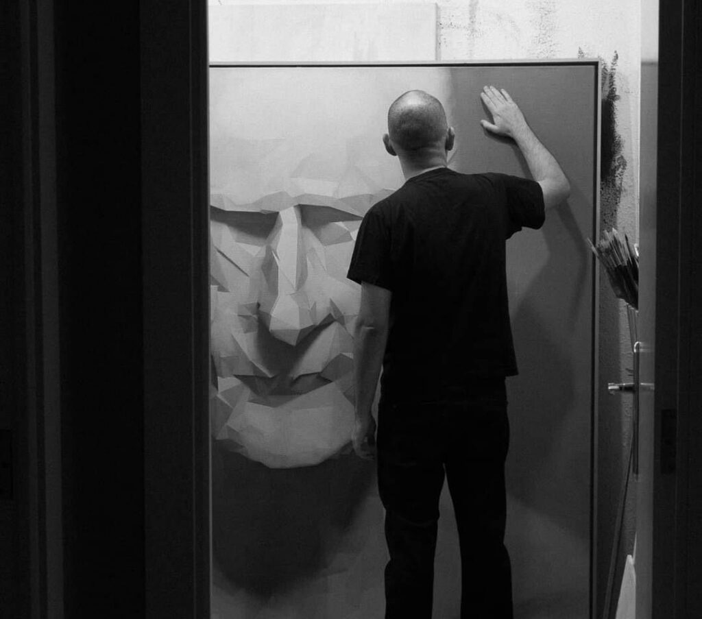 Back view of a man in a black shirt standing in a doorway, touching a geometric wall sculpture in an art studio.