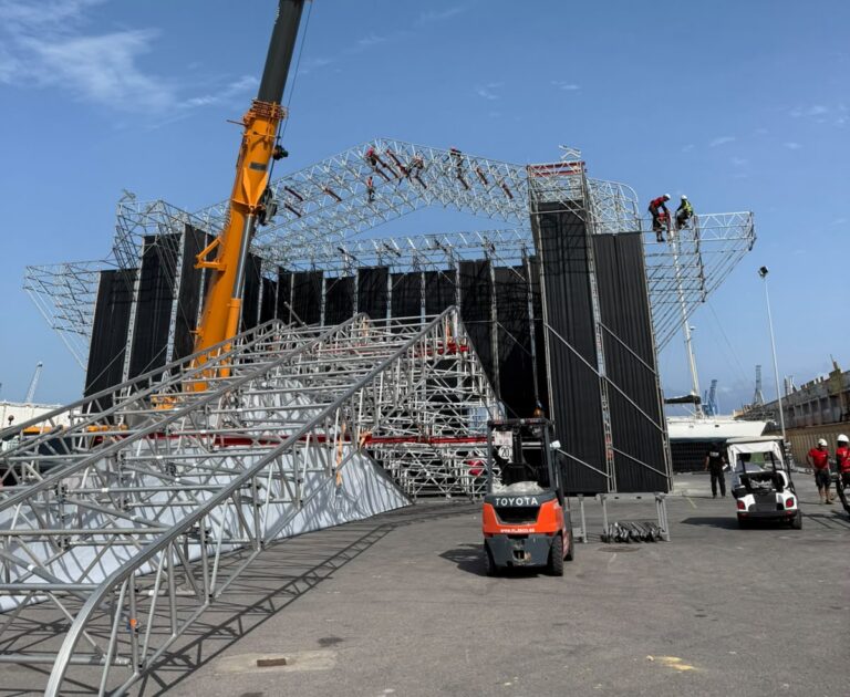 Crane lifting equipment over a large steel framework under construction, with workers on scaffolding against a blue sky.