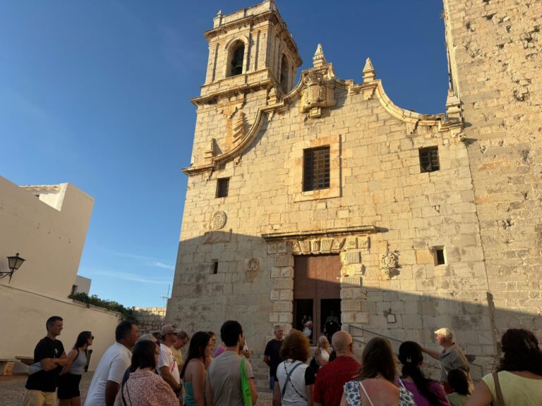 Stone church facade with arched doorway and bell tower, crowd gathered outside on a sunny day.
