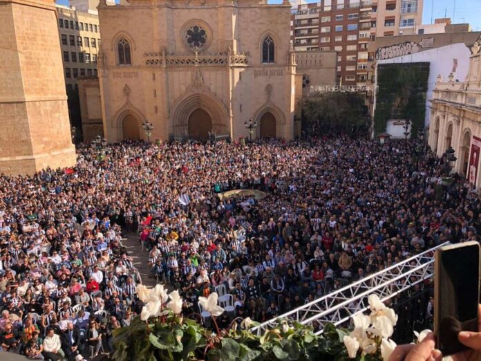 Crowd fills a plaza in front of a historic church, viewed from above, with a stage and metal truss in the foreground and city buildings in the background.