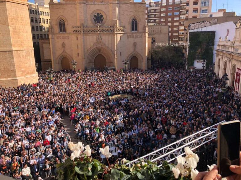 Crowd fills a plaza in front of a historic church, viewed from above, with a stage and metal truss in the foreground and city buildings in the background.