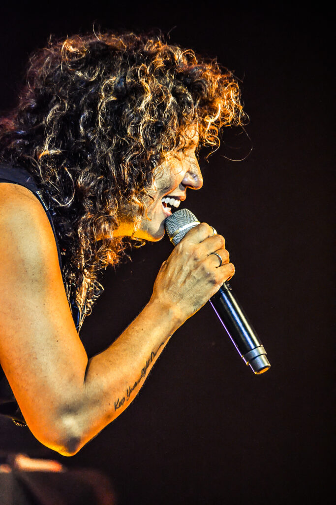 Closer view of a performer with curly hair singing into a handheld microphone on stage, side profile.