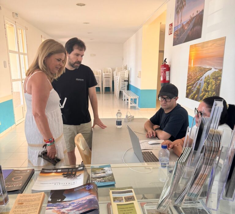 Two visitors stand at a glass brochure counter talking with two staff seated behind laptops in a bright, tiled lobby with magazines and displays on the counter.