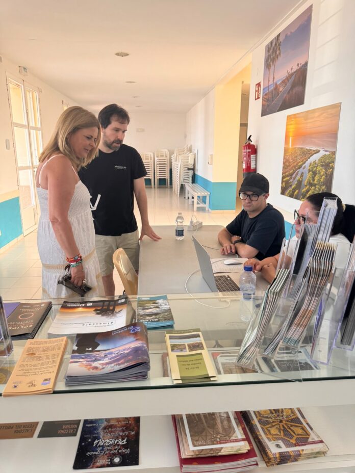 Woman in white dress and a man stand at a glass-counter reception, talking with two staff members seated behind the brochure-filled desk in a bright room.