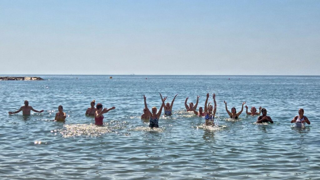 Group of people in waist-deep water at the beach, arms raised as they play in the calm sea on a sunny day.