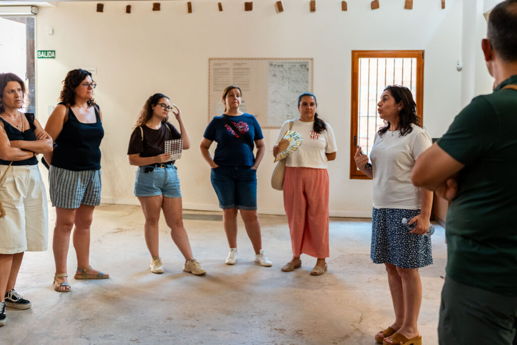 Group of adults standing in a circle inside a bright room, listening to a woman speak on the right.